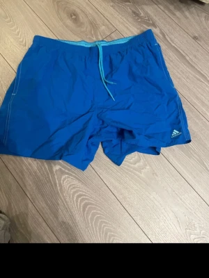 Blå badbyxor från Adidas - Snygga blå badbyxor från Adidas med elastisk midja och dragsko i ljusblått. Klassisk Adidas-logga på vänster ben. Tillverkade i snabbtorkande material, perfekta för stranden eller poolen. Enkel och sportig design.