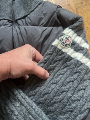 Grå pufferjacka från Moncler - Snygg grå pufferjacka från Moncler med stickade ärmar i kabelstickat mönster och vita ränder. Jackan har två dragkedjefickor fram, hög krage och ikonisk Moncler-logga på ärmen. Perfekt för kalla vinterdagar och riktigt trendig look.