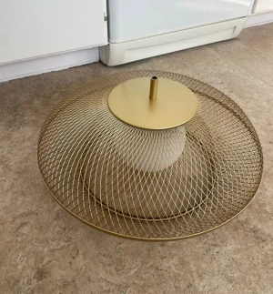 Guldig lampskärm i metallnät - Säljer en rund lampskärm i guldigt metallnät med modern design. Skärmen har ett nätmönster och en vit klotformad insats i mitten där glödlampan placeras. Perfekt för att ge rummet en stilren och trendig känsla.