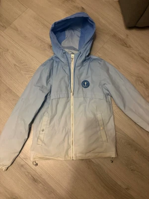 Blåvit vindjacka med huva - Snygg vindjacka i blått och vitt med huva och dragkedja framtill. Jackan har två fickor med knappar, ett runt blått märke på bröstet och coolt blått tryck på ryggen. Tillverkad i lätt material, perfekt för blåsiga dagar.