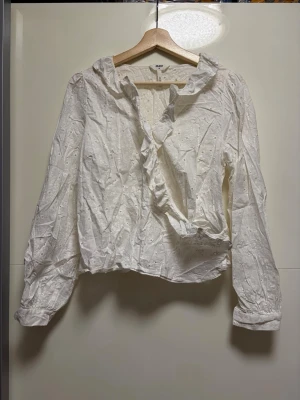 Vit blus med volang från Object - Söt vit blus från Object med volangdetalj längs den djupa v-ringningen. Blusen har ett broderat hålmönster och långa ärmar med knapp vid ärmslut. Perfekt att styla med jeans eller kjol för en trendig look.