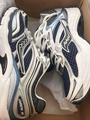 Saucony Omni 9 blåvita sneakers - Snygga Saucony Omni 9 sneakers i blått, vitt och svart med meshpaneler och detaljer i silver. Skorna har en sportig retrodesign, vita snören och en robust sula för extra komfort. Perfekta för dig som gillar klassiska löparvibbar och streetstyle.