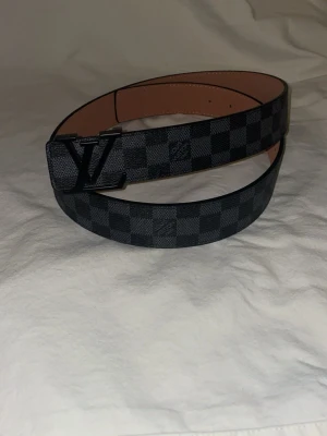 Louis Vuitton rutigt skärp svart/grå - Snyggt Louis Vuitton skärp i svart och grått med klassiskt rutigt Damier-mönster och stor LV-logga på spännet. Skärpet är gjort i skinn och har en lyxig känsla, perfekt för att lyfta vilken outfit som helst.