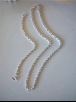 Flätat halsband och armband i äkta silver 925 - Säljer ett stilrent halsband  och armband i äkta silver 925 med flätad kedja och karbinlås. Halsbandet har en snygg, glänsande finish och en liten bricka med 925-stämpel vid låset. Perfekt för dig som gillar klassiska accessoarer med en modern twist.