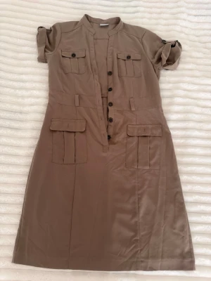 Beige skjortklänning med fickor - Stilren beige skjortklänning med korta ärmar och knappar framtill. Klänningen har flera fickor med lock både på bröstet och höfterna samt fina knappdetaljer. Materialet känns mjukt och har en lätt glans. Perfekt för en avslappnad men ändå chic look.