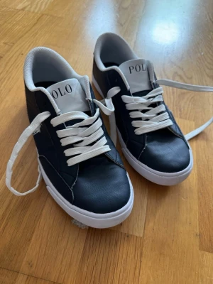 Svarta Polo Ralph Lauren sneakers - Snygga mörkblåa  sneakers från Polo Ralph Lauren med vita detaljer och vita skosnören. Klassisk låg modell med logga på sidan och märkesnamn på plösen. Ovandelen är i slätt material och insidan är fodrad i vitt tyg. Perfekta för en stilren och sportig look.