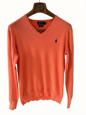 Korallfärgad stickad tröja Polo Ralph Lauren - Snygg korallfärgad stickad tröja från Polo Ralph Lauren med V-ringning och blå broderad logga på bröstet. Tröjan har långa ärmar och ribbade muddar vid ärmslut och nederkant. Perfekt för dig som gillar klassisk stil.