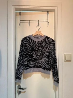 Grå zebramönstrad stickad tröja - Säljer en fluffig stickad tröja i grått med svart zebramönster. Tröjan har rund halsringning, långa ärmar och ribbade muddar vid ärmslut och nederkant. Perfekt för dig som vill ha en trendig och mysig look.