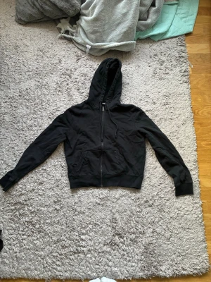 Svart zip hoodie med huva - En klassisk svart hoodie med dragkedja framtill och stor ficka på magen. Tröjan har huva med snören och ribbade muddar vid ärmslut och nederkant. Perfekt för en chill och avslappnad stil.