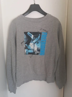 Grå sweatshirt med blått tryck - Grå sweatshirt från Lindex i storlek 158–164. Mjuk och bekväm modell med lång ärm, avslappnad passform och snyggt grafiskt tryck fram. Använd men i fint skick utan hål eller större skador. Perfekt till vardags och enkel att matcha med jeans eller mjukisbyxor.