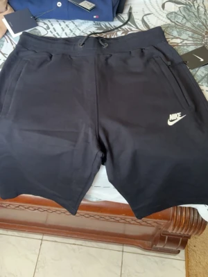 Svarta Nike shorts med snörning - Snygga svarta shorts från Nike med klassisk logga på vänster ben. Shortsen har snörning i midjan, två sidofickor med dragkedja och är tillverkade i mjuk bomullsmix. Perfekta för träning eller chill dagar.