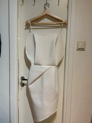 Vit figurnära klänning - Stilren vit klänning asymmetrisk nederkant. Figurnära passform och dragkedja i ryggen för enkel påklädning. Perfekt för dig som vill ha en minimalistisk och modern look.
