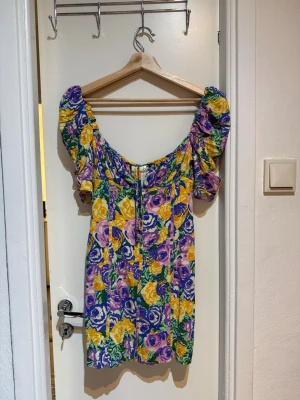Blommig kortklänning från Zara - Färgglad kortklänning från Zara med blommigt mönster i lila, gul, grön och vit. Klänningen har puffiga korta ärmar, markerad midja och dragkedja bak. Perfekt för sommarens alla tillfällen!