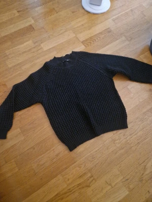 Svart stickad tröja från 157 - Mysig svart stickad tröja från 157 med grovstickad struktur och ribbade muddar vid hals, ärmslut och nederkant. Perfekt för lager på lager och enkel att matcha till olika outfits.
