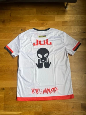 JUL-t shirt  - Unik vit t-shirt med färgglada grafiska tryck och alienmotiv på ryggen. Svarta och röda detaljer vid krage och ärmslut. Framsidan har texten 'D'or et de platine' och coola färginslag i gult, orange och svart. Perfekt för dig som gillar streetwear och statement-plagg. Storleken är XS men passar även S. 