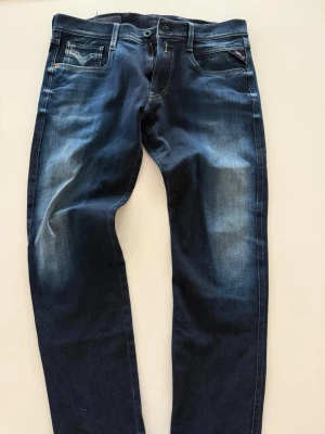 Mörkblå Replay jeans med slitningar - Snygga mörkblå jeans från Replay med diskreta slitningar och tvättade detaljer på låren. Klassisk femficksmodell med raka ben och knappgylf. Märkesdetalj på fickan och kontrastsömmar. Perfekta för en trendig och avslappnad stil.