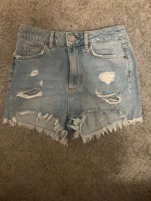 Ljusa jeansshorts med slitningar - Säljer ett par ljusblå jeansshorts med råa, fransiga kanter och flera slitningar både fram och bak. Klassisk femficksmodell med knapp och dragkedja. Perfekta för en avslappnad och trendig sommarstil.