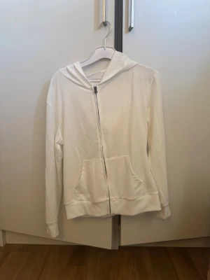 Vit zip hoodie med fickor - Snygg vit hoodie med dragkedja framtill och två stora fickor. Hoodien har en klassisk huva och ribbade muddar vid ärmar och nederkant. Perfekt för en avslappnad stil. Materialet känns mjukt och bekvämt, troligen bomull eller bomullsblandning.