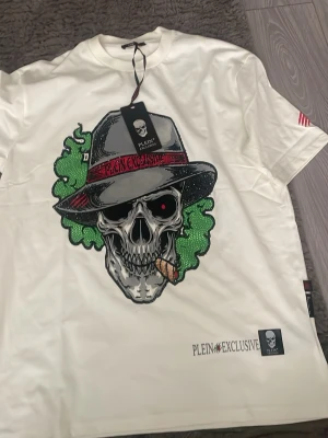 Vit t-shirt från Plein Exclusive oversize - Snygg vit t-shirt från Plein Exclusive med stort tryck framtill av en tecknad figur med hatt och grönt hår. T-shirten har korta ärmar och rund hals. Märkeslogga och textdetaljer finns både fram och på etiketten. Tillverkad i mjuk bomull.