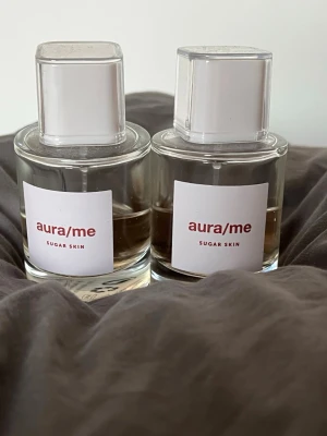 Aura/Me Sugar Skin parfym - Två flaskor Aura/Me Sugar Skin parfym med minimalistisk design. Flaskorna är genomskinliga med vita fyrkantiga lock och etiketter med röd text. Doften är söt och modern, perfekt för dig som gillar fräscha och sockriga toner. 
