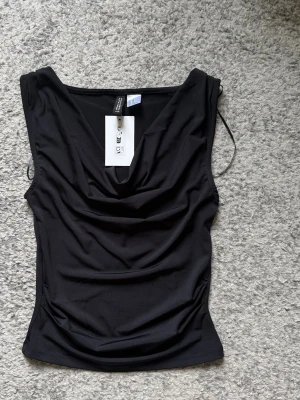 Svart ärmlös topp från H&M - Snygg svart ärmlös topp med draperad v-ringning. Tillverkad i mjuk polyester med en figurnära passform. Perfekt att styla med jeans eller kjol för en trendig look.