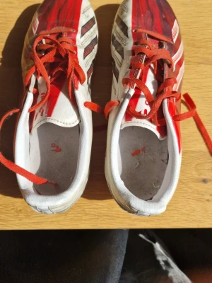 Adidas vita och röda fotbollsskor - Säljer ett par Adidas fotbollsskor i vitt och rött med svarta detaljer och röda skosnören. Skorna har klassisk dobbsula för gräsplan och är tillverkade i syntetmaterial. Adidas-logga på sidan och hälen. Perfekta för dig som vill sticka ut på planen.