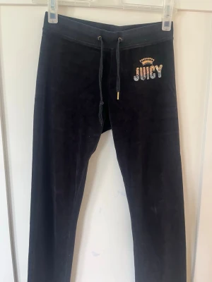 Svarta velourbyxor från Juicy Couture - Mörka, mjuka velourbyxor från Juicy Couture med dragsko i midjan och glittrande 'JUICY'-logga framtill. Byxorna har raka ben och två fickor med knappar bak. Perfekta för en chill och trendig look.
