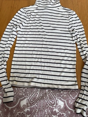 Randig polotopp från H&M Basic - Vit och svart randig polotopp från H&M Basic. Toppen är ribbad med lång ärm och har en klassisk polokrage. Perfekt att styla med jeans eller kjol för en snygg och enkel look.