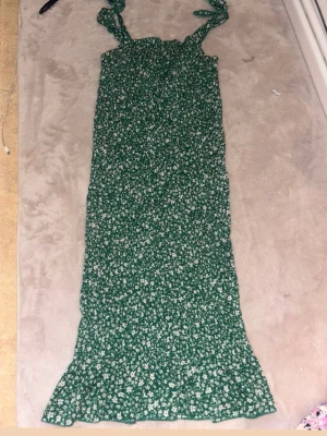 Grön blommig långklänning med knyt - Grön långklänning med små vita blommor och knytbara axelband. Klänningen har en smickrande rak passform och volangkant nertill. Perfekt för sommarens alla tillfällen.