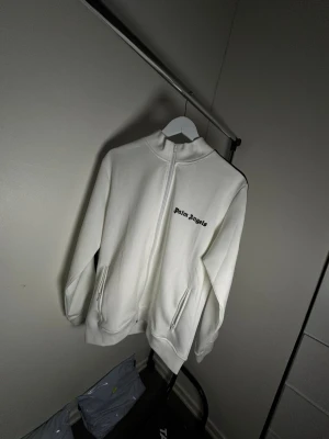 Vit zip hoodie från Palm Angels - Säljer en vit hoodie från Palm Angels med hel dragkedja och svarta ränder längs ärmarna. Hoodien har hög krage, två fickor med dragkedja och Palm Angels-logga på bröstet. Tillverkad i mjukt material som känns skönt mot huden.