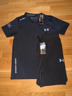 Svart träningsset från Under Armour - Snyggt svart träningsset från Under Armour med t-shirt och shorts. Båda plaggen har färgglada loggor och tryck, samt bekväm passform. Perfekt för gymmet eller löprundan. Materialet är lätt och andas bra.