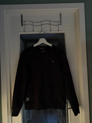 Mörkblå sweatshirt från Morris - Mörblå sweatshirt med rund hals och diskret broderad logga på bröstet. Tröjan har ribbade muddar vid ärmslut och nederkant samt en liten patch nertill. Perfekt för en avslappnad och stilren look.