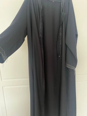 Svart abaya med broderade detaljer - Säljer en svart abaya med vida ärmar och tunna broderade detaljer längs kanterna och vid ärmsluten. Tyget är tunt och faller snyggt, perfekt för dig som gillar en modest och elegant stil. Knappar funkar ej