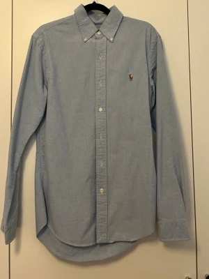 Ljusblå skjorta från Ralph Lauren - Klassisk ljusblå skjorta från Ralph Lauren med button-down krage och broderad logga på bröstet. Skjortan har lång ärm, normal passform och knäppning framtill. Perfekt till jeans eller chinos för en clean och stilren look.