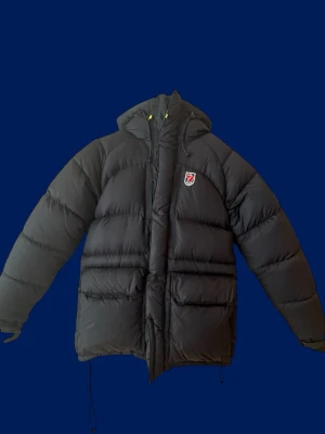 Mörkblå dunjacka från Fjällräven - Snygg mörkblå dunjacka från Fjällräven med huva och dragsko. Jackan har två stora framfickor och klassisk Fjällräven-logga på bröstet. ..Perfekt för kalla vinterdagar tack vare det tjocka dunfodret och den höga kragen. Passar-S . 