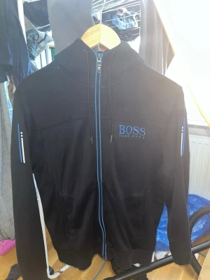 Svart hoodie från Hugo Boss - Snygg svart hoodie från Hugo Boss med blå detaljer och dragkedja framtill. Märket är broderat i blått på bröstet. Hoodien har huva och vita samt blå ränder längs ärmarna. Perfekt för en stilren och sportig look.