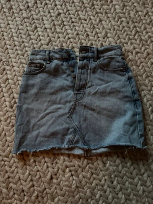 Ljusblå jeanskjol med rå kant - Snygg ljusblå jeanskjol i kort modell med rå, fransig nederkant och klassiska fickor fram och bak. Kjolen har knappstängning framtill och bälteshällor. Perfekt för en avslappnad och trendig look. Går att klippa kortare utan att bli förstörd
