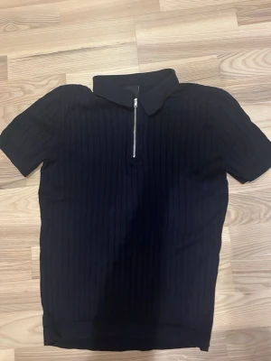 Svart  polotopp med dragkedja - Svart ribbad polotopp med kort ärm och halv dragkedja framtill. Toppen har en klassisk krage och är tillverkad i ett stretchigt material som sitter snyggt på kroppen. Perfekt för en stilren och modern look.