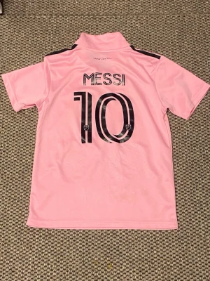 Inter Miami Messi matchtröja rosa - Säljer en rosa Inter Miami matchtröja från Adidas med Messi och nummer 10 på ryggen. Tröjan har svarta detaljer, klubbmärke på bröstet och sponsorlogga framtill. Tillverkad i lätt och ventilerande material, perfekt för fotboll eller träning.