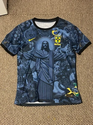Brasilien tröja  - Snygg blå matchtröja från Nike med Brasil-logga och gult swoosh. Tröjan har ett detaljerat grafiskt tryck med Kristusstatyn och brasilianska motiv. Svarta muddar vid ärmar och hals. Perfekt för dig som älskar fotboll och vill sticka ut på planen.