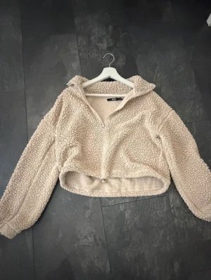 Beige teddytröja med halv dragkedja - Mysig beige teddytröja med halv dragkedja och hög krage. Tröjan har en avslappnad passform, långa ärmar och är tillverkad i ett fluffigt, mjukt material som känns riktigt skönt mot huden.