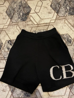 Svarta stickade shorts med CB-detalj - Svarta stickade shorts med bred resår i midjan och stora fickor både fram och bak. På vänster ben finns en stor vit CB-logga som ger en cool kontrast. Perfekta för en avslappnad och trendig look.