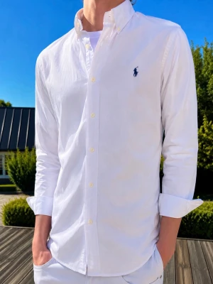 Vit skjorta från Ralph Lauren - (MODELLEN ÄR 185CM OCH VÄGER 76KG)Klassisk vit skjorta från Ralph Lauren med button-down krage och broderad logga på bröstet. Skjortan har lång ärm, knappar framtill och är tillverkad i bomull. Perfekt för en clean och stilren look.