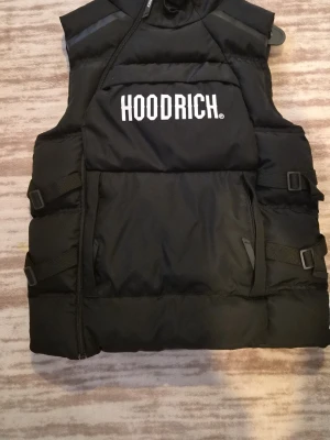 Svart dunväst från Hoodrich - Svart dunväst från Hoodrich med stor vit logga på bröstet. Västen har quiltad design, justerbara band i sidorna och dragkedja framtill. Perfekt för lager-på-lager och streetwear-stil.