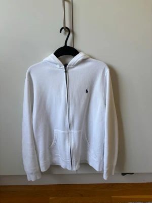 Vit hoodie från Polo Ralph Lauren - Snygg vit hoodie från Polo Ralph Lauren med dragkedja framtill och klassisk huva. Tröjan har fickor på magen och den ikoniska lilla loggan broderad på bröstet. Tillverkad i mjukt bomullsmaterial som känns skönt mot huden. Perfekt för en clean och stilren look. Luvan är lite skrynklig inuti på grund av att den är precis ny tvättad