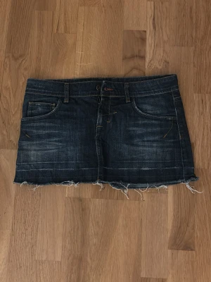 Mörkblå jeanskjol med rå kant - Snygg mörkblå jeanskjol i kort modell med slitna detaljer och rå, fransig kant nedtill. Klassisk femficksdesign och bälteshällor. Perfekt för en avslappnad och trendig look. Stolek 38 men skulle säga att den sitter som en S på mig