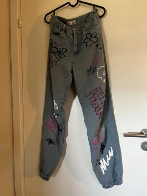 Baggy jeans med graffiti och blommor - Unika baggy jeans i ljusblå denim med graffiti-motiv, blommor och text i rosa och svart. Resår vid benslut och hög midja ger en cool och avslappnad vibe. Perfekta för dig som vill sticka ut med din stil.