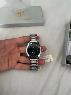 Silverfärgad klocka från Pablo Raez - Stilren klocka från Pablo Raez med svart urtavla och datumvisning. Armbandet är i blankt silverfärgat metall och har en klassisk länkdesign. Perfekt accessoar för dig som gillar en tidlös och elegant look.