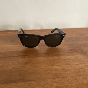 Svarta Ray-Ban Wayfarer solglasögon - Klassiska svarta Wayfarer solglasögon från Ray-Ban med mörka glas och ikonisk form. Snygg och tidlös design med Ray-Ban-logga på sidan. Perfekta för soliga dagar och ger en cool vibe till din outfit. Endast använda 2 gånger. Perfekt skick.