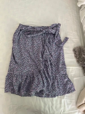 Ljuslila blommig omlottkjol - Söt ljuslila omlottkjol med blommigt mönster och volangkant. Kjolen har knytband i midjan och är i storlek xs från Only